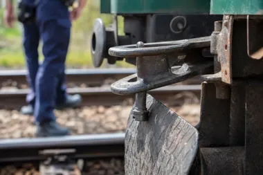 Zbliżenie na metalowy zderzak i sprzęg kolejowy na tle torów oraz stojącego w tle umundurowanego funkcjonariusza.