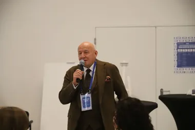 Mężczyzna w brązowym garniturze przemawia do publiczności przez mikrofon podczas konferencji w sali konferencyjnej.